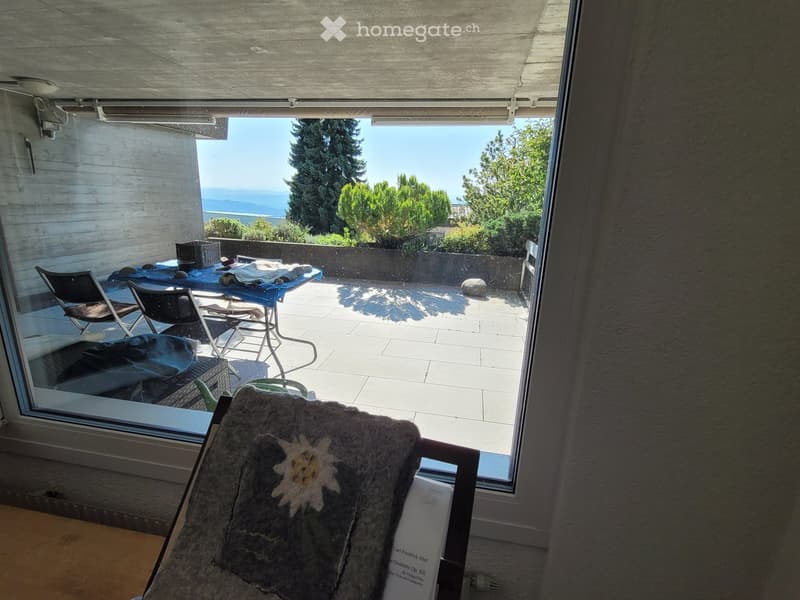 Balcony with tiled floor, outdoor furniture including chairs and a table, covered by a retractable awning, overlooking a scenic view of the sea and greenery.