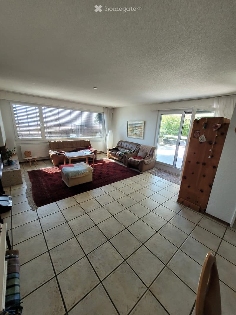 Bright living room with tiled floor, large windows, and sliding glass doors leading to a balcony. Features a brown sofa, coffee table, ottoman, and a small wooden chair.