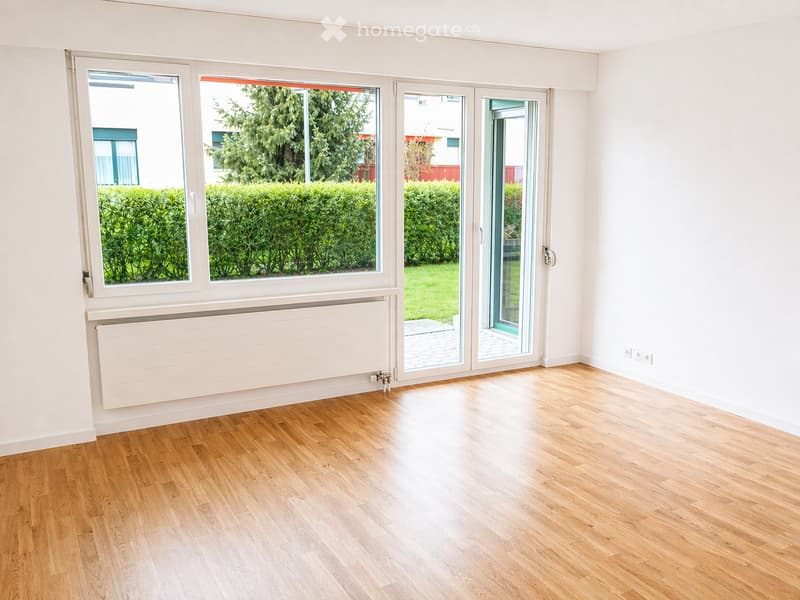 Empty room with wooden flooring, white walls, large windows, and a sliding glass door leading to a garden.