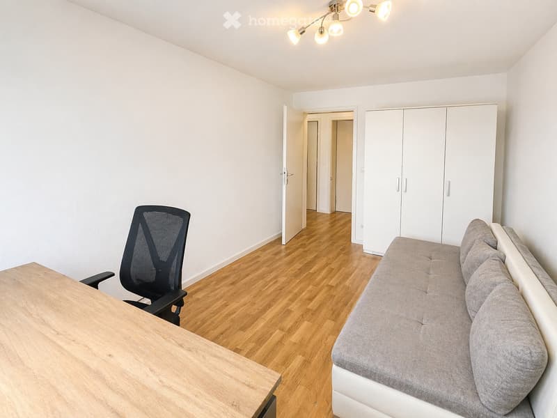 A modern, well-lit room with a grey sofa, a black office chair, and a wooden table. The room features light wooden flooring and white walls, with a large built-in wardrobe and a ceiling light fixture.