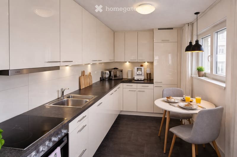 Modern kitchen with white cabinets, black countertops, stainless steel sink, stove, microwave, coffee maker, and a small dining area with a round table and chairs.