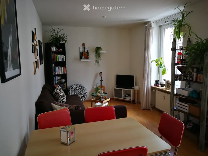 A cozy living room with a sofa, coffee table, dining table with chairs, bookshelf, television, and various decorative items including plants and a guitar.