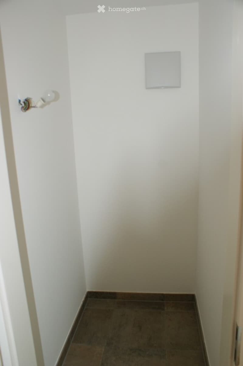 Empty room with white walls and dark tiled floor, featuring a light switch and a light fixture on the wall.