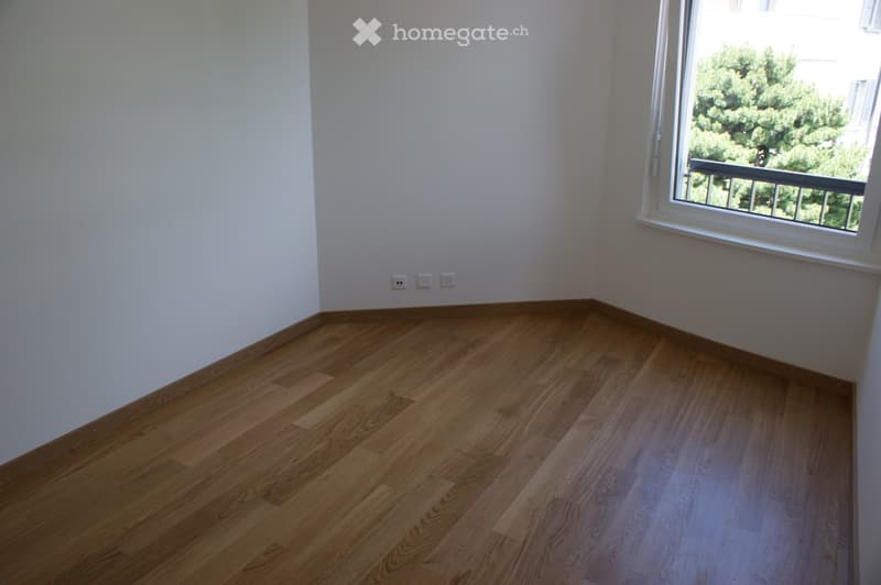 Empty room with light wooden flooring and white walls, featuring a large window with a view of greenery and a balcony outside.