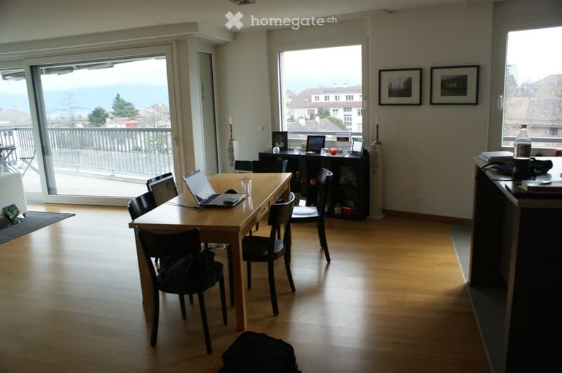 Bright room with wooden floor, large windows, balcony access, table with laptop, chairs, shelf, and decorative items.