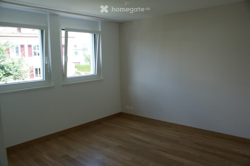 Empty room with wooden flooring, white walls, and two large windows with white frames, allowing natural light to enter.