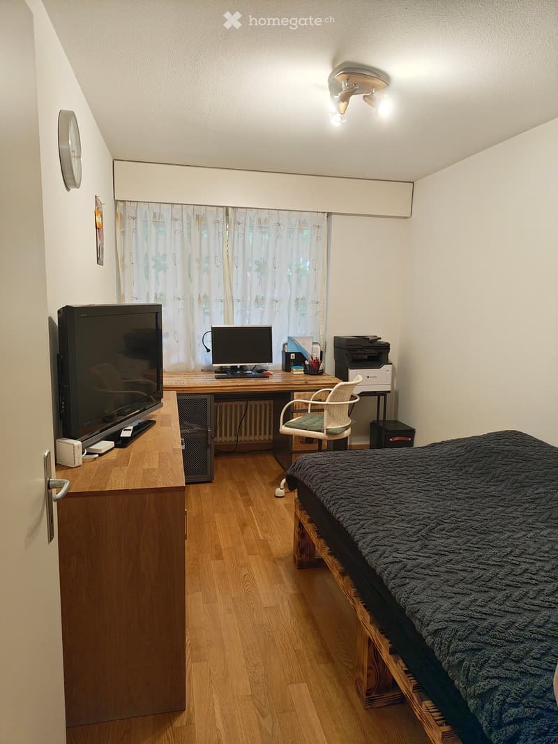 Cozy bedroom with a wooden floor, a bed with a dark blanket, a desk with a computer, a chair, a television, and a printer. The room has white walls, a window with floral curtains, and a ceiling light fixture.