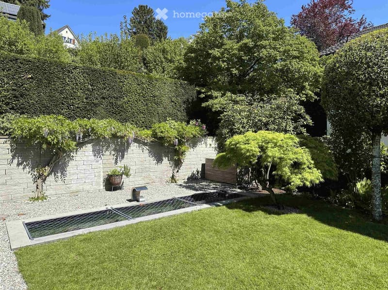 Well-maintained garden with a lawn, gravel pathways, a small water feature, and various trees and shrubs. The area is enclosed by a brick wall and features a modern design with clean lines and a serene atmosphere.