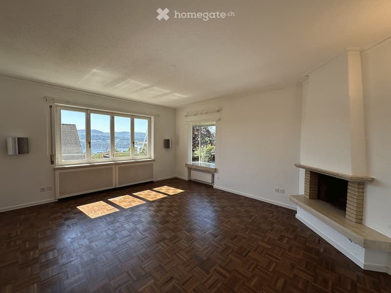 Empty room with wooden flooring, large windows offering scenic views, and a fireplace with a stone surround.
