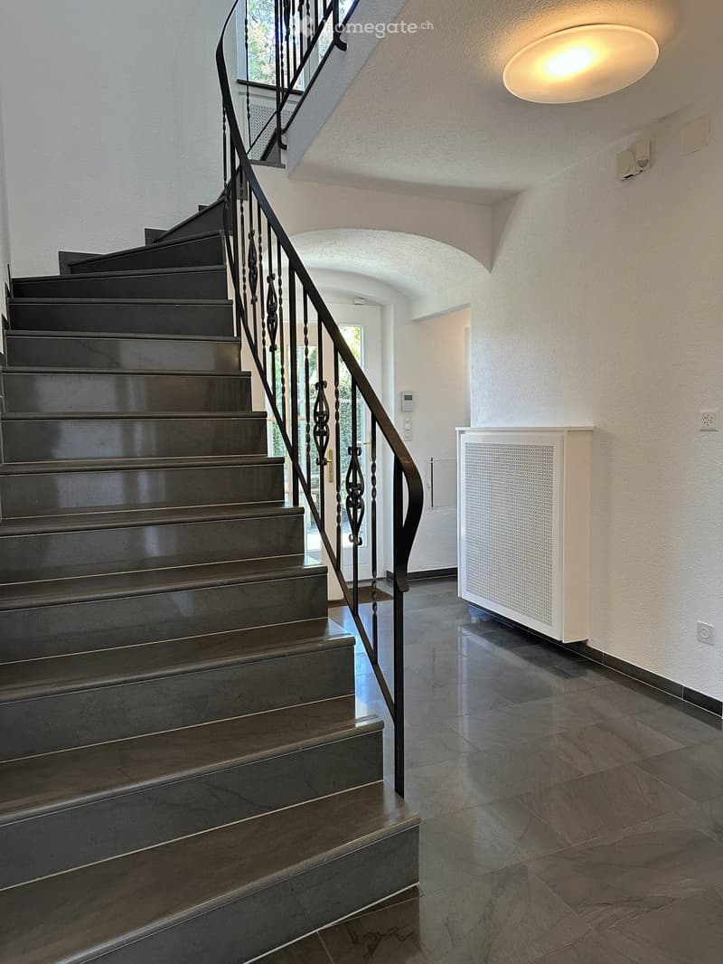 Staircase with dark wooden steps and a black wrought-iron railing, leading to an open area with a white wall and a round ceiling light.