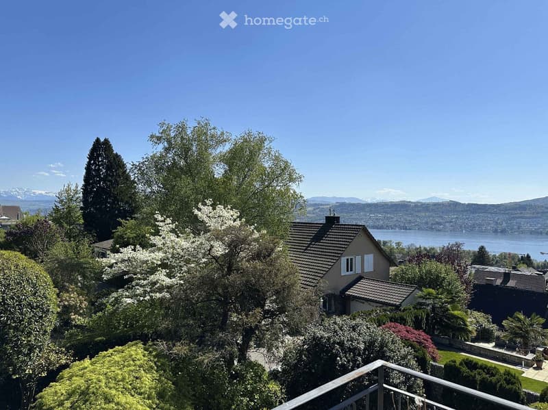 A scenic outdoor view from a balcony or terrace, showing a house surrounded by lush greenery, with a body of water and distant mountains in the background.
