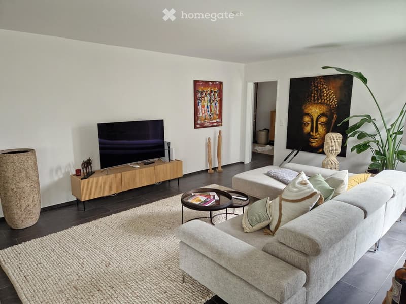 Modern living room with a grey sectional sofa, coffee table, TV, sideboard, and decorative plant. The space features a large, open layout with white walls and dark wooden flooring.