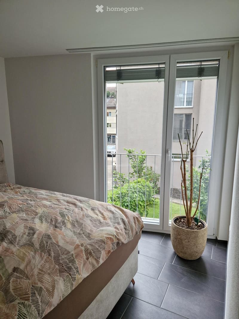 Bedroom with a bed covered in a patterned duvet, a large potted plant near the balcony entrance, and a view of a green outdoor area through large windows and a sliding door.
