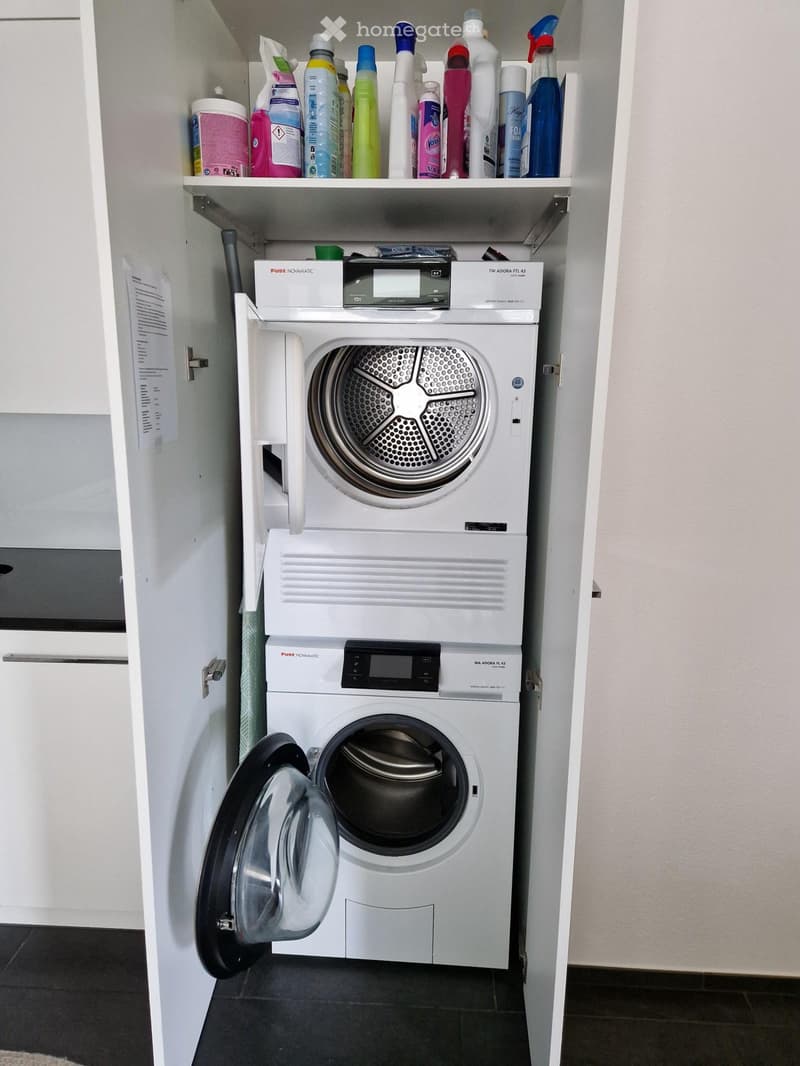 Compact laundry area with a stacked washer and dryer unit, open shelving with cleaning supplies, and white cabinetry.