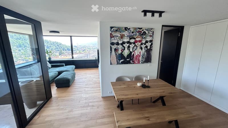 Modern living and dining area with wooden flooring, large windows offering a scenic view, a teal sofa, wooden dining table with chairs, and a colorful abstract painting on the wall.