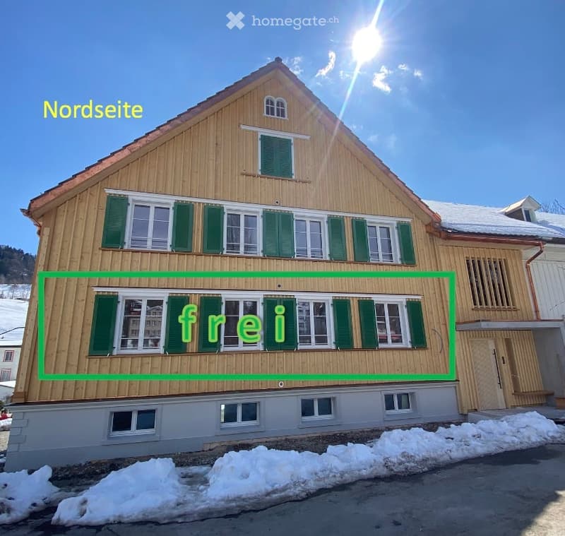Traditional wooden house with green shutters, multiple windows, and a pitched roof, surrounded by snow.