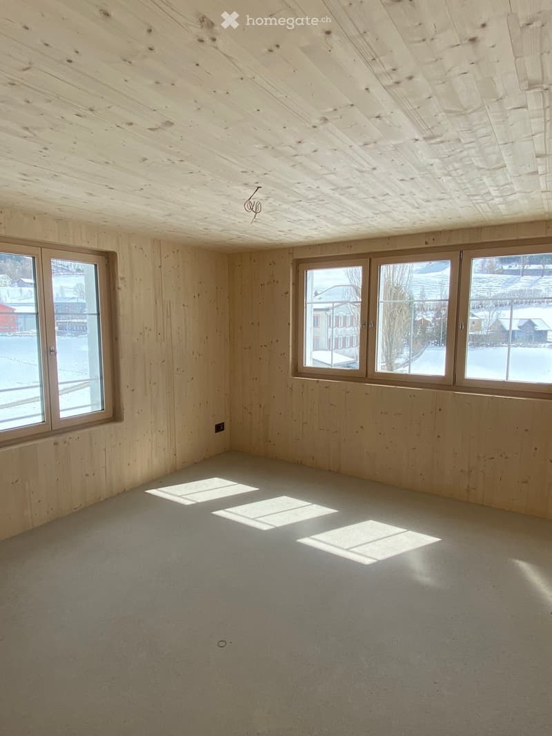 Empty room with wooden walls and ceiling, large windows, and a carpeted floor. Natural light streams in from the windows, casting shadows on the floor.