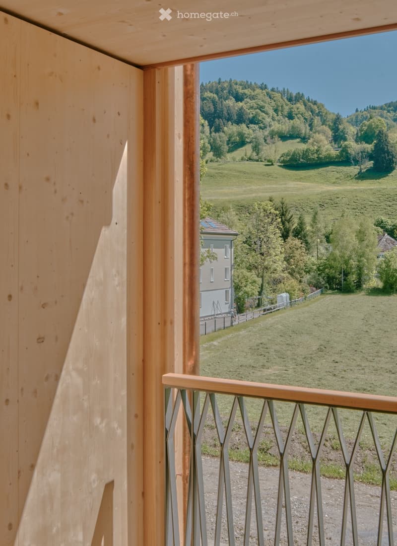 Balcony with wooden railing and metal balusters, overlooking a green valley with hills and a house in the distance.
