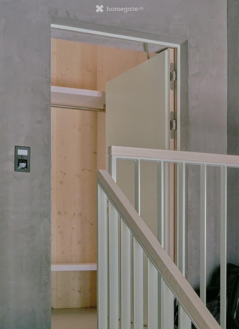 A modern hallway with a staircase leading upwards, featuring a wooden railing and a partially open closet door revealing shelves inside.