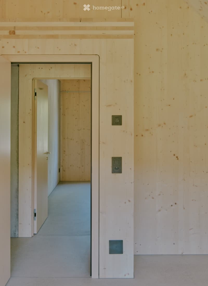 Minimalist hallway with light wood paneling, concrete floor, and a single open door leading to another space.