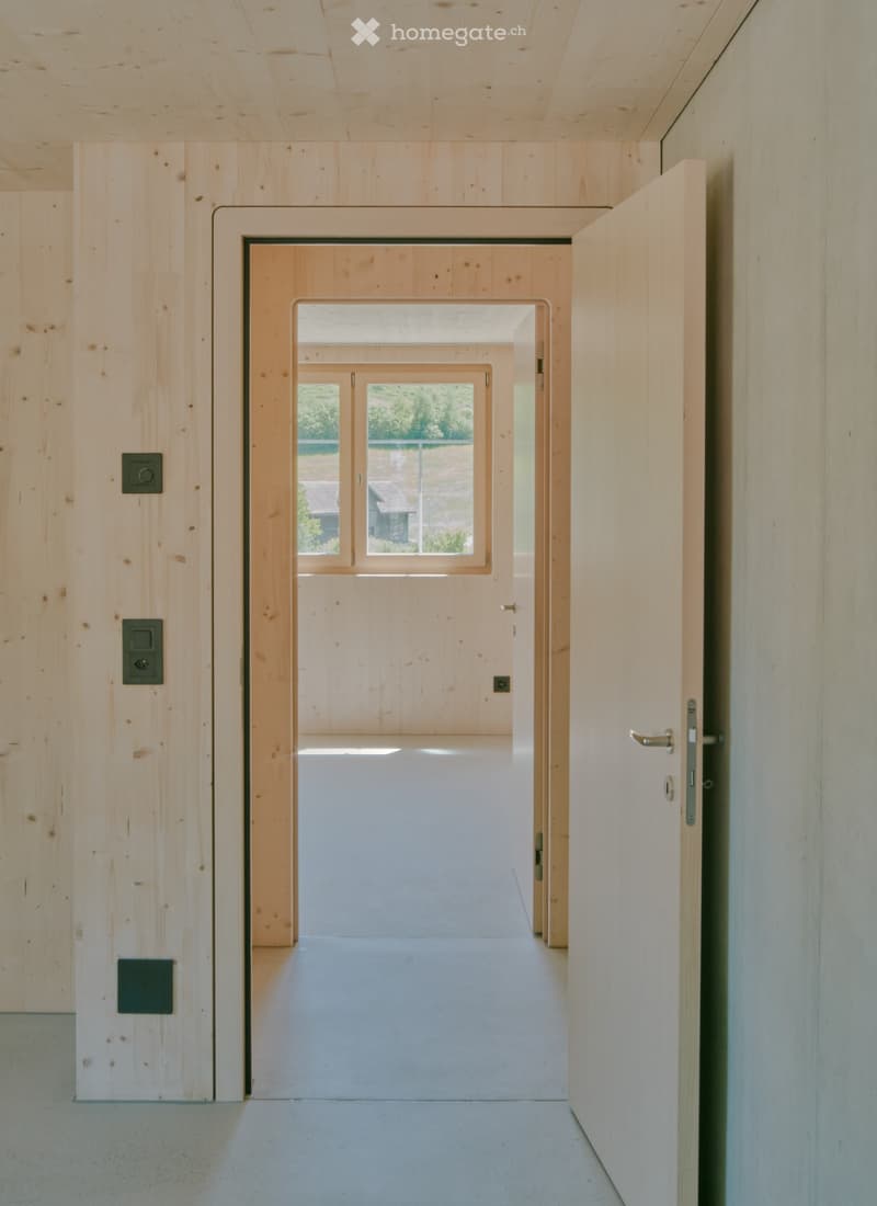 Minimalist wooden hallway with open doorways leading to a bright, airy space. Natural light streams in from the far end, revealing a view of greenery and a building outside.