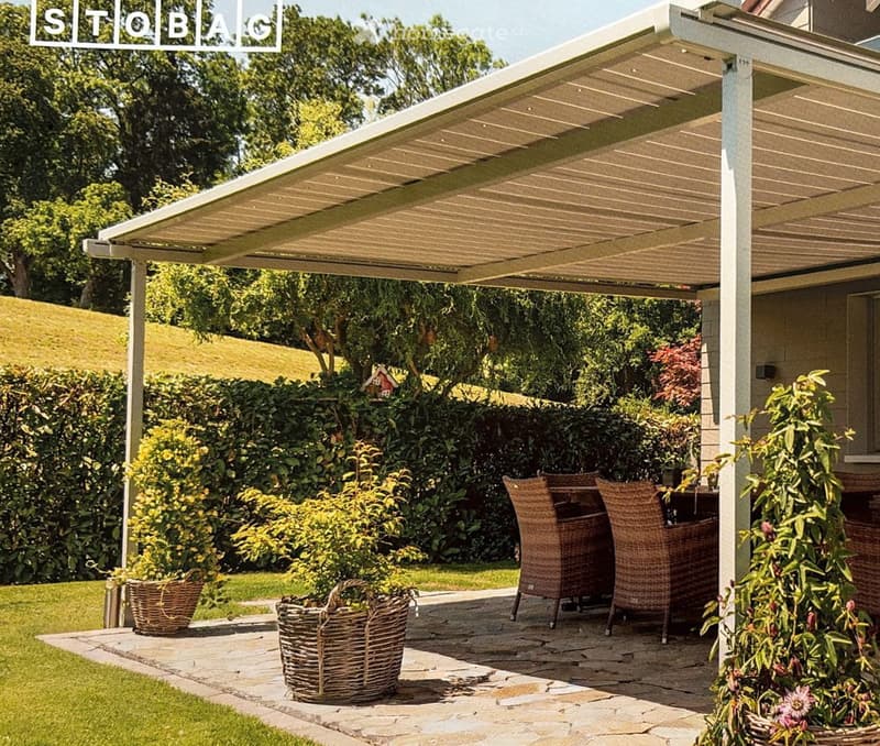 A covered outdoor patio with wicker chairs and tables, surrounded by lush greenery and potted plants. The area features a stone-paved floor and a modern pergola roof.
