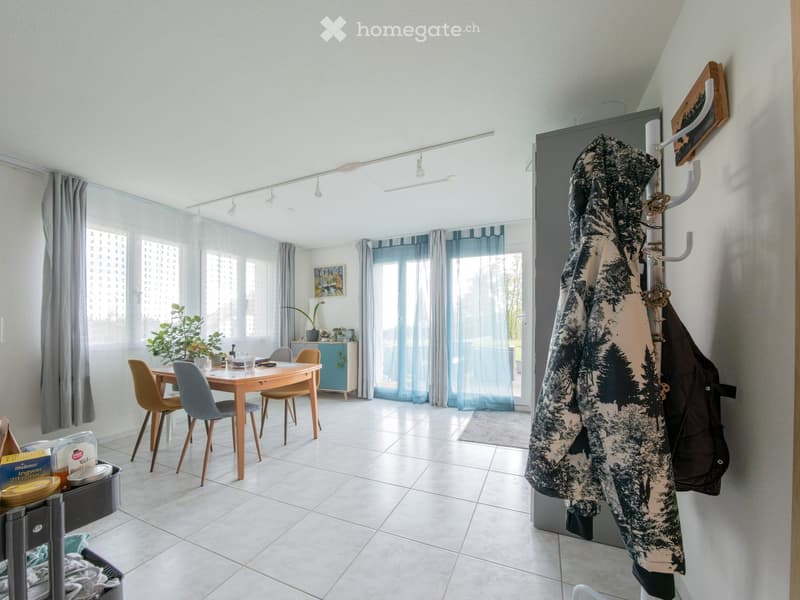 Bright, modern room with white tiled flooring, large windows with sheer curtains, a wooden dining table with four chairs, a small cabinet, and a coat rack with jackets.
