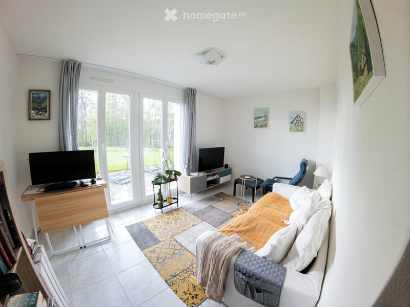 Bright living room with large windows and sliding doors leading to a patio. Features a white sofa with a throw blanket, a wooden coffee table, a TV stand, a bookshelf, and a small round table with a chair.