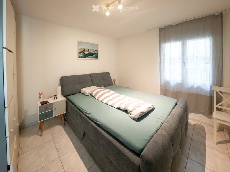 Cozy bedroom with a grey upholstered bed, light blue bedding, a nightstand with a lamp, a white chair, and a painting on the wall.