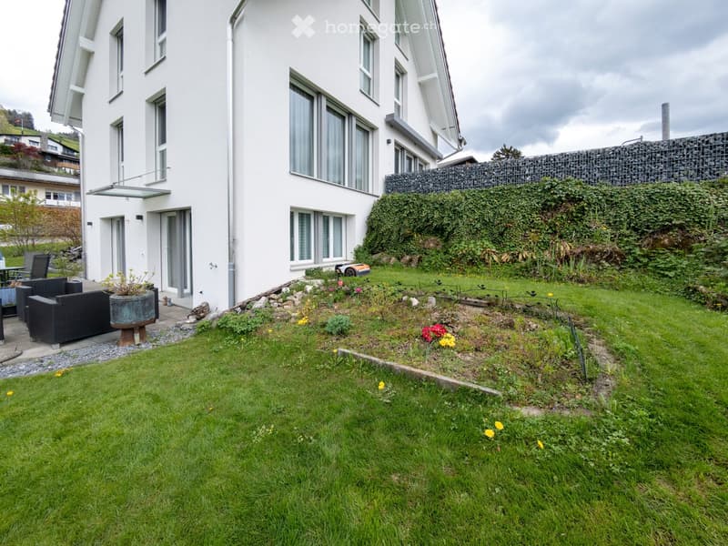 Well-maintained garden with green lawn, flower beds, and a small seating area with black wicker chairs and a table near the house entrance.