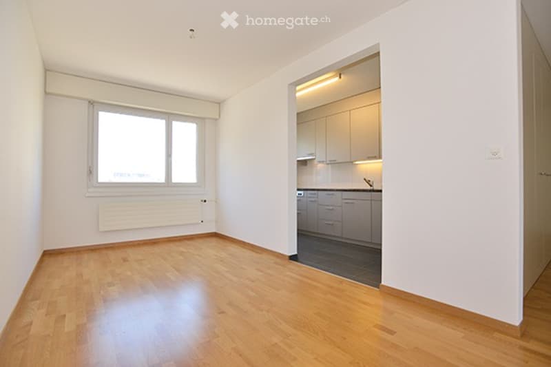 Empty room with wooden flooring, white walls, and a large window. Adjacent kitchen area with cabinets and appliances is visible through an open doorway.