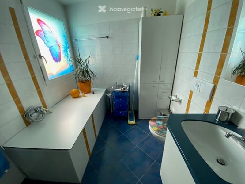 Bathroom with white tiled walls, blue tiled floor, sink, toilet, cabinet, and a small plant on the windowsill.