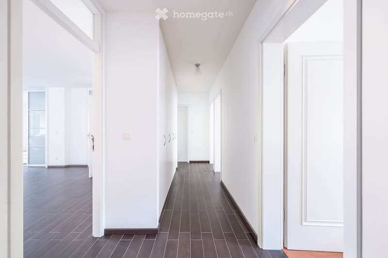 Corridor / Hallway. A long, narrow hallway with white walls, dark wooden flooring, and several doors on both sides. The hallway is well-lit with recessed lighting and has a modern, minimalist design.