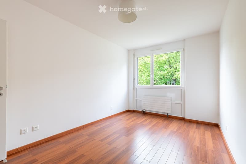 Unfurnished Room. Empty room with white walls, wooden flooring, and a window with a view of greenery. Includes a radiator and a ceiling light fixture.