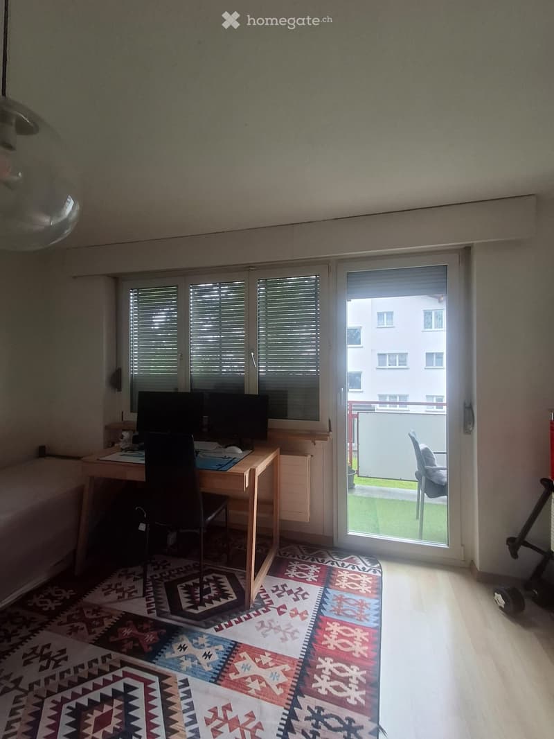 Bright room with wooden flooring, colorful patterned rug, desk with computer and chair, large window with blinds, and sliding glass door leading to a balcony.