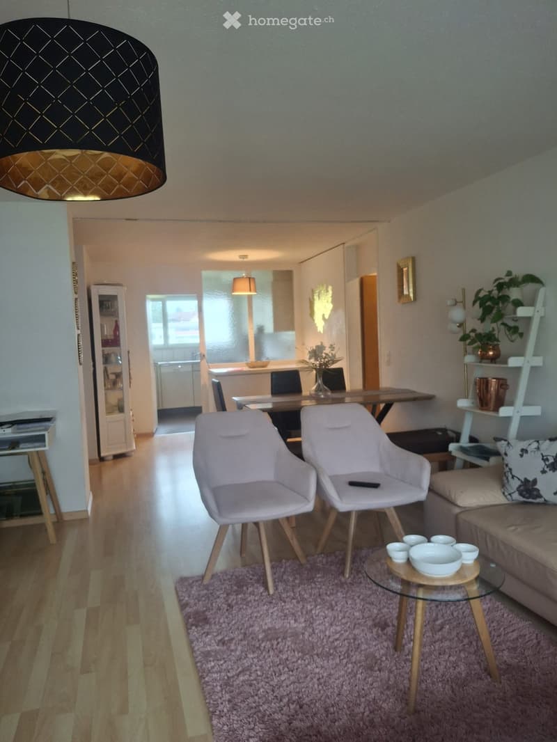 Modern living room with light-colored furniture, including a sofa, two armchairs, a coffee table, and a dining area with a table and chairs. The space features a purple rug, wooden flooring, and decorative plants.