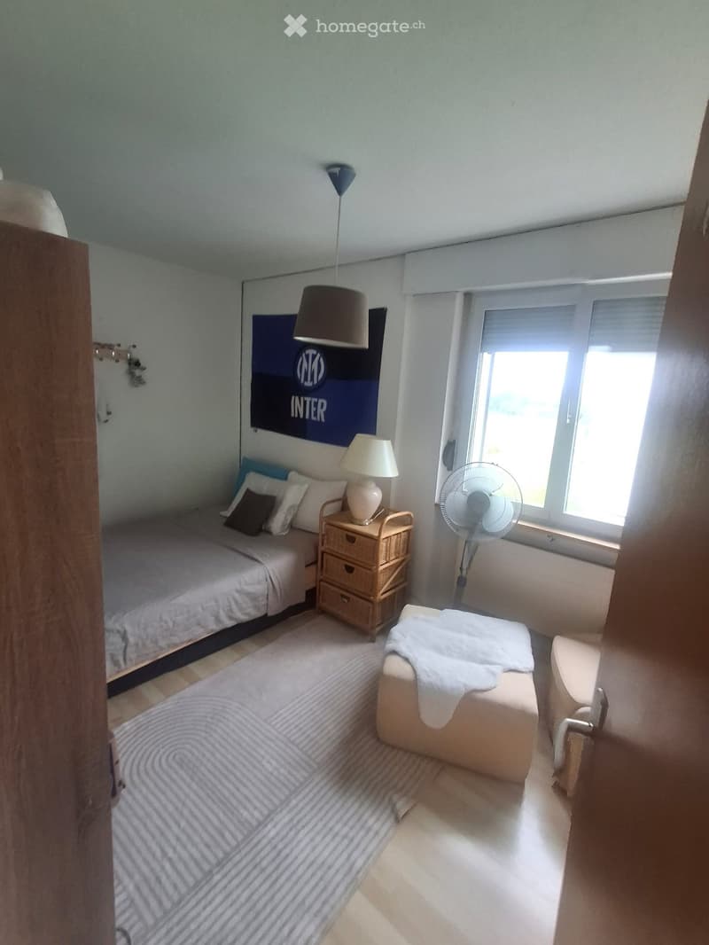 Cozy bedroom with a single bed, nightstand, lamp, fan, and a small seating area with a chair and stool.