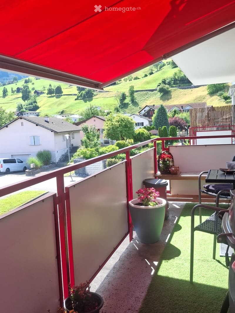 Balcony with a scenic view of green hills and houses, featuring a red awning, potted plants, a chair, and a table.