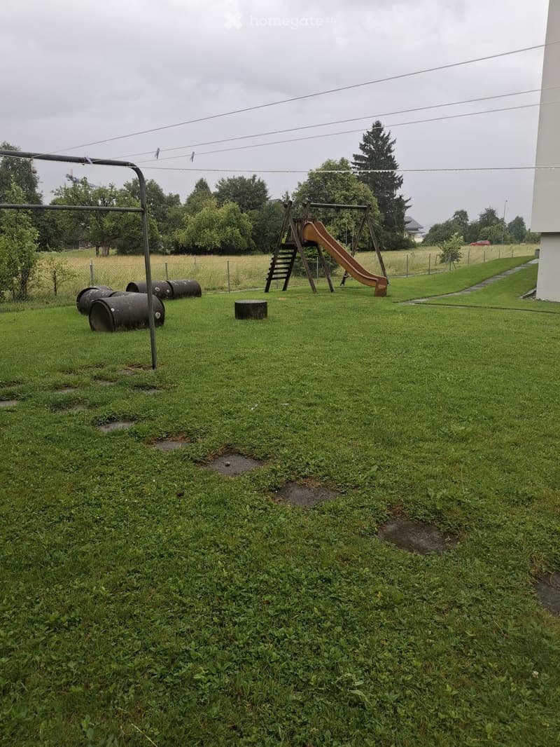 A grassy outdoor area with a wooden slide, swing set, and several large black barrels. The area is bordered by a fence and trees, with a pathway leading to a building on the right.