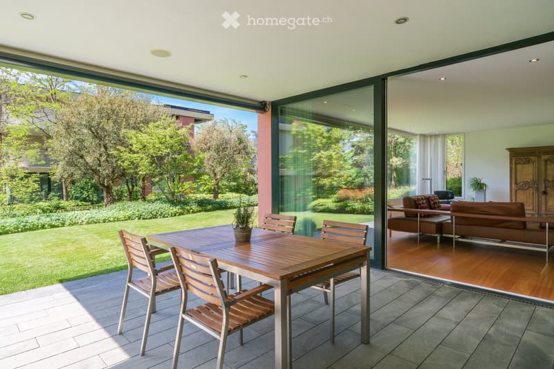 A modern, spacious balcony with a wooden table and chairs, overlooking a well-maintained garden with trees and greenery. The balcony features large glass doors leading to an indoor living area with a brown leather sofa and ottoman.
