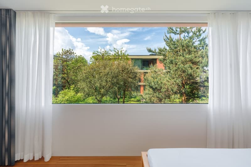 Empty room with wooden flooring, white walls, large window with white curtains, and a view of trees and a building outside.