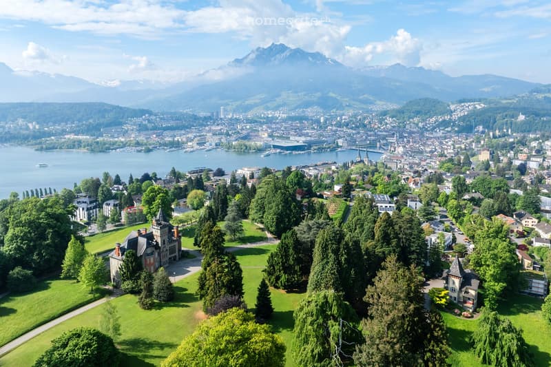 Aerial view of a city with a lake and surrounding mountains, featuring a mix of residential and commercial buildings, green spaces, and a waterfront area.