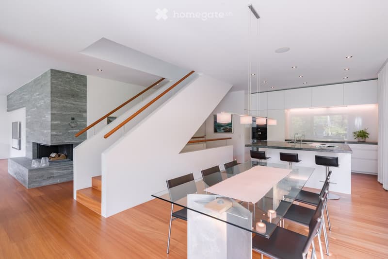 Open-concept living and dining area with a glass dining table, modern kitchen island, bar stools, and a fireplace. The space features wooden flooring, pendant lighting, and a staircase leading to an upper level.