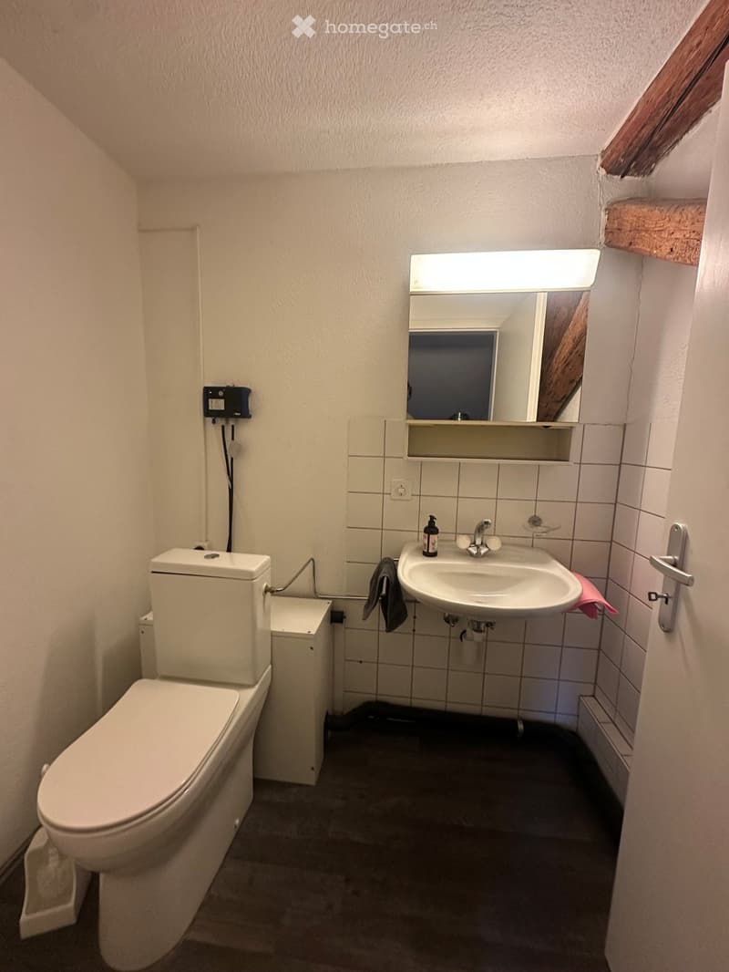 Compact bathroom with a toilet, sink, mirror, and soap dispenser. The walls are tiled, and the floor is dark.