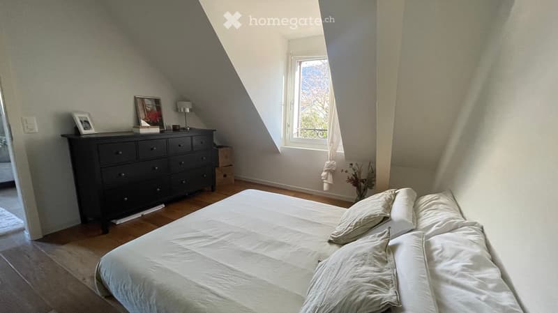 Cozy attic bedroom with wooden floors, slanted ceilings, large window, bed, dressers, lamp, and picture frame.