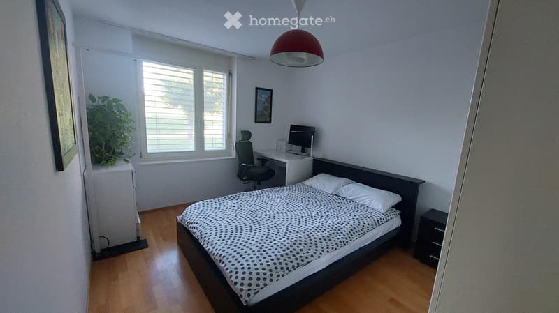 A well-lit bedroom with a double bed, desk, chair, and nightstand. The room features wooden flooring, large windows with blinds, and a hanging red lamp.