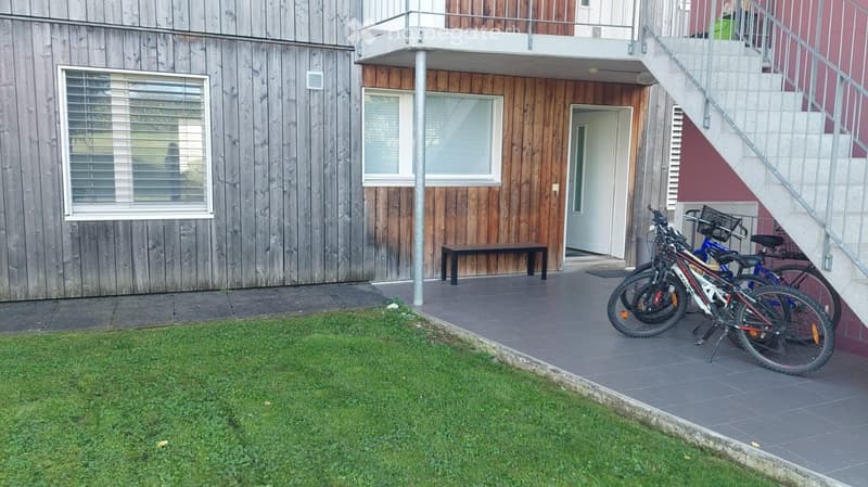 Outdoor area with a wooden building, a small grassy patch, a bench, and several bicycles parked near a staircase leading to an upper level.