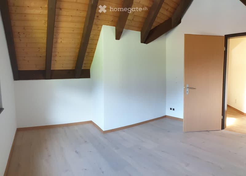 Empty room with light wooden flooring, white walls, and exposed wooden beams on the ceiling. A single door leads to another space.
