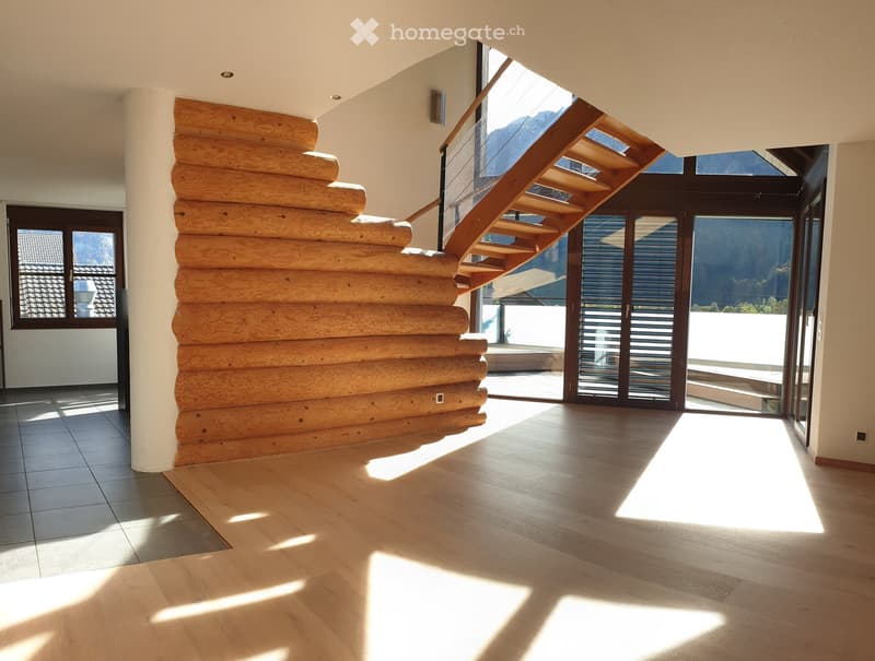 Bright, modern hallway with wooden flooring, large windows, and a staircase leading upwards. Natural light streams in, creating a spacious and airy feel.