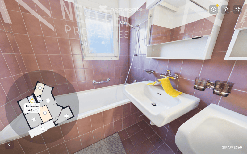 Bathroom with a bathtub, sink, mirror, and a yellow towel on the sink. The walls are tiled in a warm, reddish-brown color, and there is a window providing natural light.